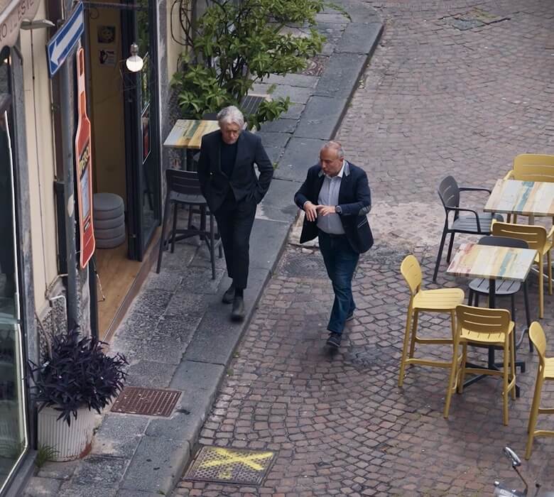 Researchers walk on a cobblestone street past a cafe in Italy, discussing plastic pollution.